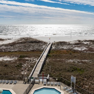 Expansive beachfront view with a wooden walkway leading to the beach, framed by lush dunes and sparkling water