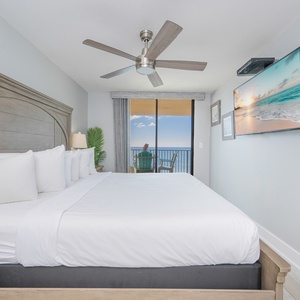 Master bedroom featuring a king bed, ceiling fan, and a balcony with beach front views, complemented by a mounted TV