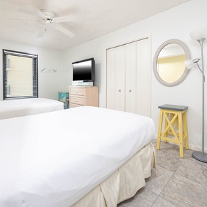 Bright and airy guest room featuring two full beds, a TV, and cheerful accents like a yellow stool and a floor lamp