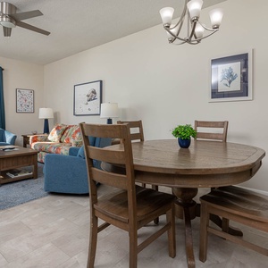 Bright living area features a floral sofa, blue armchairs, and a dining table, with expansive views of the beach beyond
