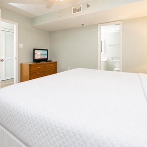 The bedroom features a large king bed, a wooden dresser with a TV, and an adjoining bathroom visible through an open door