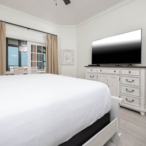 The master bedroom features a large TV, light-colored decor, and a view of the beachfront through elegant sliding doors