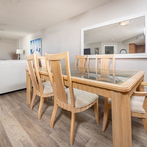 A wooden dining table with glass top and beige chairs is set against a backdrop of a bright living area and beach view