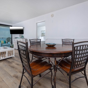 The living room features a round dining table, comfortable chairs, and a stunning beachfront view through large windows
