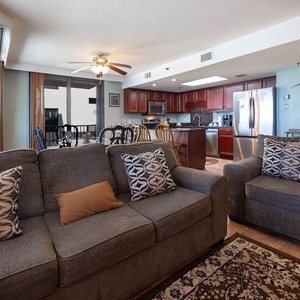 The living area features a cozy gray sofa with decorative pillows, a dining table, and the kitchen in the background