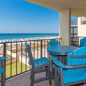 The balcony features a round table surrounded by vibrant blue chairs, offering a view of the beach and umbrellas