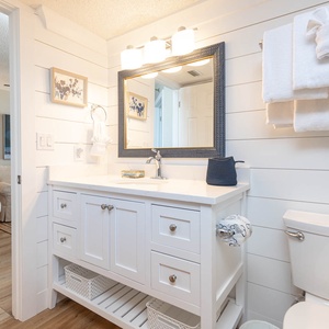 Modern bathroom featuring a white vanity, large mirror, and neatly arranged towels, with a cozy living area visible nearby