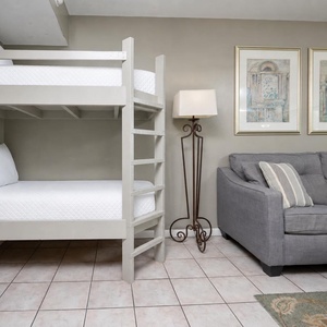 The living area features a twin bunk bed with white linens and a gray sofa adorned with striped pillows beside decorative art