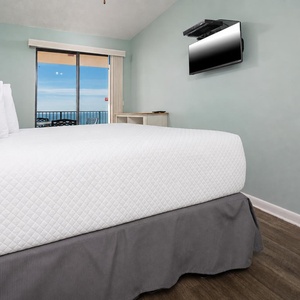 Cozy master bedroom features a plush bed, a mounted TV, and sliding glass doors leading to a balcony with a beach view