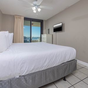 The master bedroom featuring a king bed, a mounted TV, and access to the balcony with views of the beach