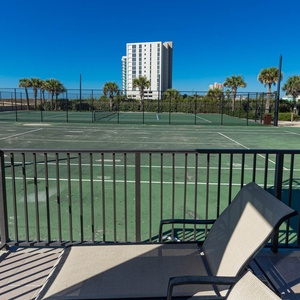 Relax on the balcony with comfortable seating, overlooking the tennis courts surrounded by lush greenery