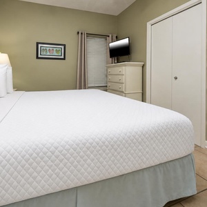 A comfortable bedroom features a plush king bed with white linens, a dresser, and a mounted TV against a soft green wall