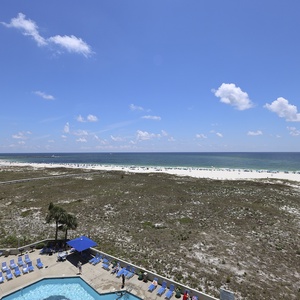 Expansive view of the beach with white sand, turquoise waters, and a pool area featuring lounge chairs and umbrellas