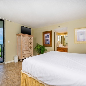 Bright master bedroom featuring a large king bed, a TV on a wooden dresser, and access to a balcony with a scenic view