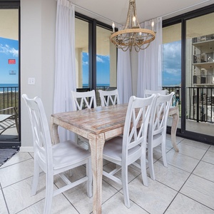 Bright dining area features a rustic wooden table surrounded by white chairs, with views of the beach through large windows