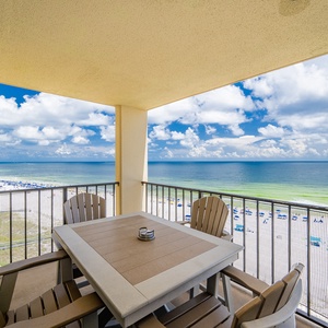 The balcony features a table and chairs with a panoramic view of the beach, showcasing vibrant blue waters and white sand