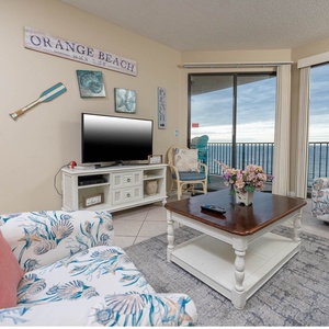 The living room features coastal-themed decor, a TV, queen sofa-sleeper, and a view of the beach through large glass doors