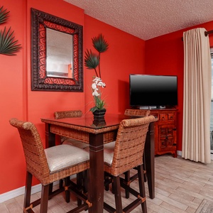 The dining area features a glass-top table with wicker chairs, a large TV, and a stunning beach view through sliding doors