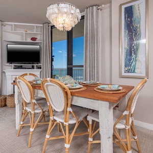The dining area features a rustic table set for four, a TV for entertainment, and a stunning beach view through large windows
