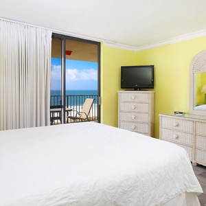 Bright yellow walls complement the master bedroom featuring a king bed, a TV, and a balcony with a stunning beach view