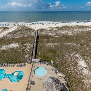 Aerial view showcases the pool area with loungers, leading to a boardwalk that connects to the beach and vibrant umbrellas