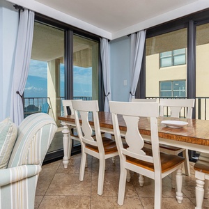 A light-filled dining area features a rustic wooden table surrounded by white chairs, with beach views