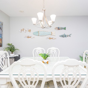 Dining area features a stylish glass-top table, white chairs, complemented by coastal wall decor and colorful accents
