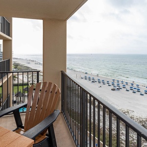 A cozy balcony features a wooden chair and table, offering a view of the beach with blue umbrellas lining the shore