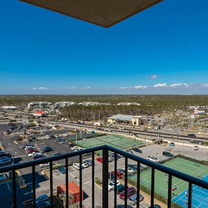 A panoramic view from the master balcony showcases nearby tennis courts, parking lots, and a vibrant blue sky