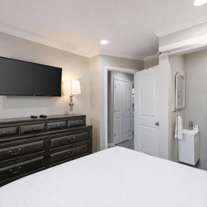 This bedroom features a sleek dresser, a mounted TV, charming lamp, and ensuite half bath creating a cozy retreat