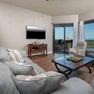 Cozy living area features a plush blue sofa with queen sleeper, a sleek TV, and access to a balcony with beach views