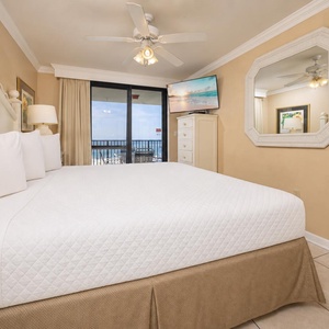 Cozy master bedroom featuring a king bed, a TV, and a balcony with views of the beach, complemented by soft, neutral decor