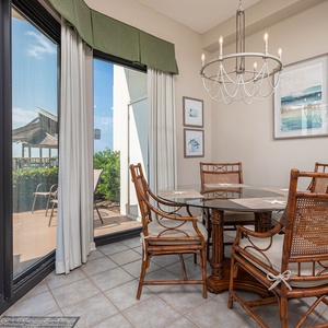 Dining area featuring a glass-top table surrounded by wicker chairs, with views of the beach visible through large windows