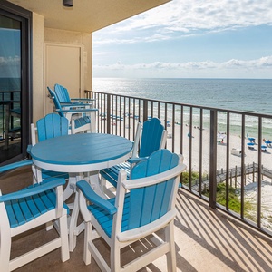 The balcony features a round table and colorful blue chairs, offering a view of the beach and umbrellas along the shore
