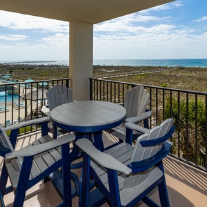Balcony features a round table and four high-backed chairs, offering a view of the beach and gentle waves beyond