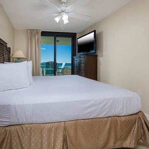 Cozy master bedroom featuring a queen bed, a TV, and access to a balcony with views of the beach and outdoor seating