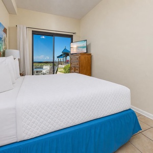 The inviting master bedroom features a plush king bed and a TV, leading to a patio and pool area with beachfront access