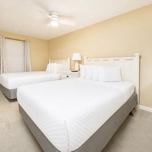 Bright and airy guest room featuring two queen beds, a ceiling fan, and a bedside table with a lamp for a restful stay