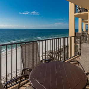 The balcony features a round table and wicker chairs, offering a serene view of the beach and turquoise waters