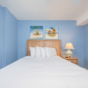 This bedroom features soothing blue walls, a comfortable bed with white linens, and vibrant beach-themed artwork