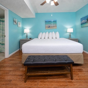 The bedroom features a king bed with white linens, a stylish black bench, and vibrant turquoise walls adorned with beach art