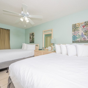This guest room features two queen beds with crisp white linens, a ceiling fan, and tropical artwork against soft green walls