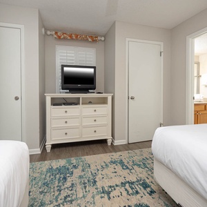 Bright guest room featuring two double beds, a dresser with a TV, and an adjoining bathroom visible through an open door