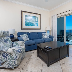 This living area features a cozy blue couch, a patterned recliner, and a stunning beach view through sliding glass doors
