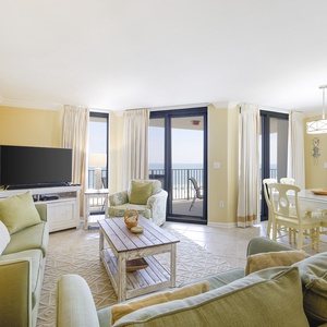 Bright living space features green sofas, a TV, and dining space with a view of the gulf through large glass doors