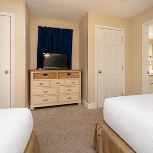 Cozy guest room features two double beds, a dresser with woven baskets, and a TV, with an adjoining bathroom visible
