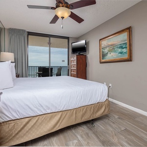 Cozy master bedroom featuring a plush king bed, a mounted TV, and sliding glass doors leading to a balcony with a beach view