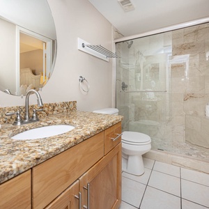 The bathroom features a granite countertop, a round mirror, and a glass-enclosed walk-in shower with elegant tile accents