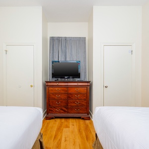 Bright and airy guest room featuring two double beds, a wooden dresser with a TV, and a doorway leading to a bathroom area