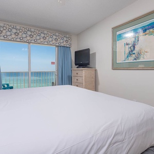 The master bedroom features a plush bed, a TV, and a stunning beachfront view through large sliding doors